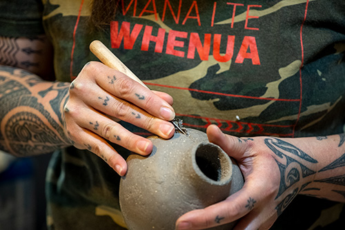 A woman's tattooed hands are holding a clay vase – she is carving shapes into it.