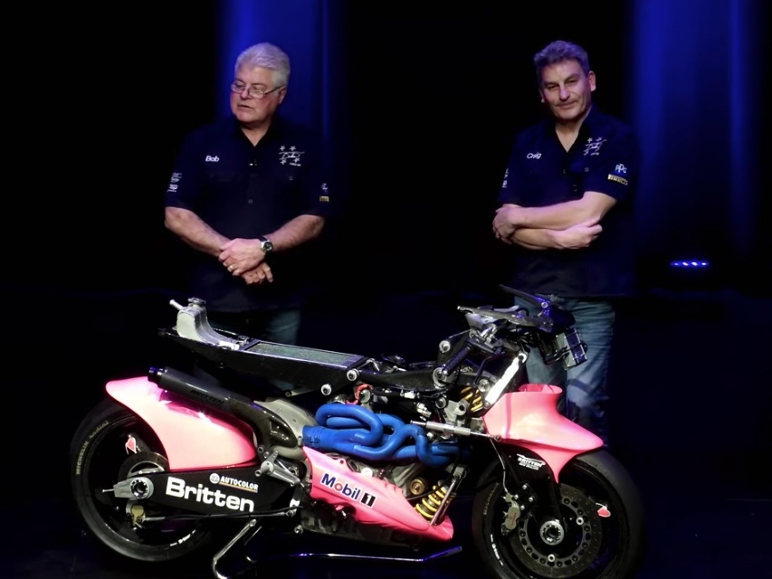Two men standing on a stage with a bright blue and pink motorbike in front of them