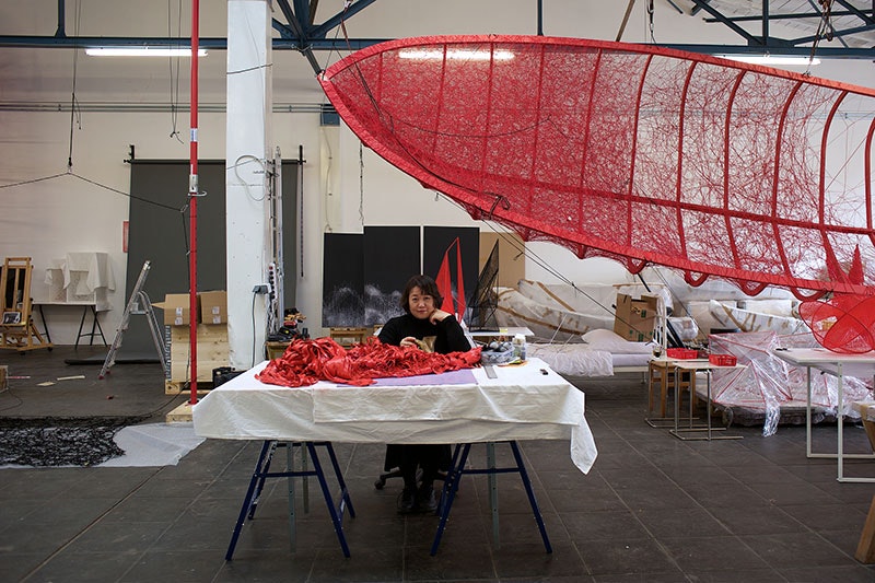 Portrait of Chiharu Shiota sitting at her desk in her studio. Above her hangs the red frame of a sailing boat frame with red wool being threaded across it