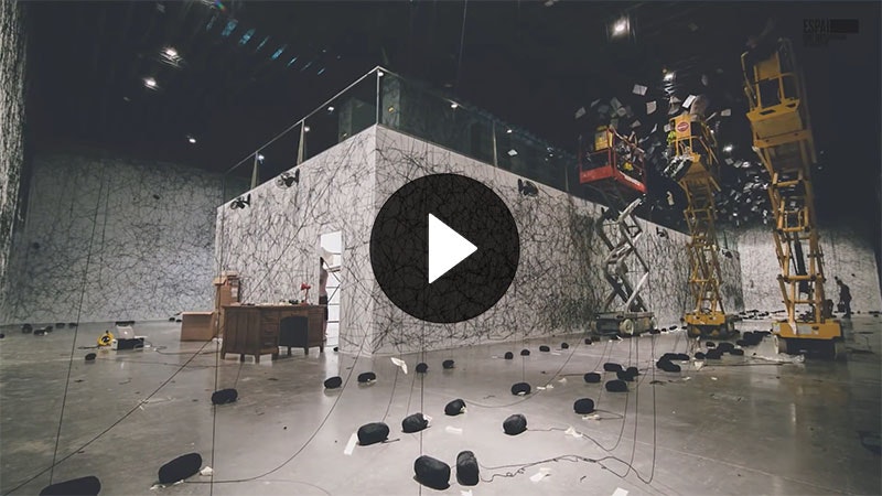 Still from a video. It shows a large pavilion in the middle of a room, three scissor lifts, and multiple strands of black wool hanging from the ceiling mid-installation