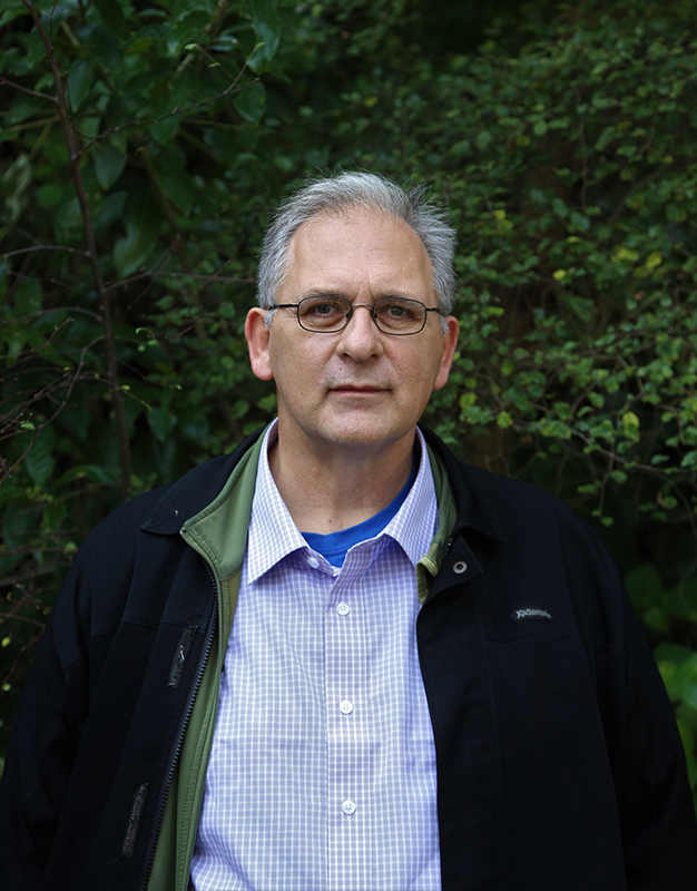 A man standing and looking at the camera. He is standing in front of a hedge