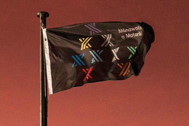 A black flag with the logo of the Matariki public holiday, consisting of nine stylised stars, flies in the early morning Wellington breeze, with an orange sunrise behind it
