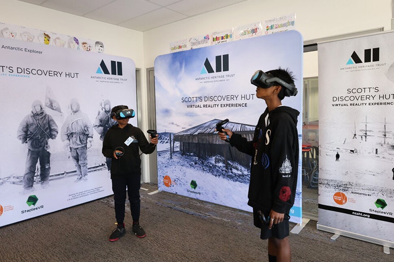 Photo courtesy New Zealand Antarctic Heritage Trust Two young boys wearng VR headsets and standing in front of three signs advertising an immersive antarctic virtual reality experience anotu Scott's Hut..