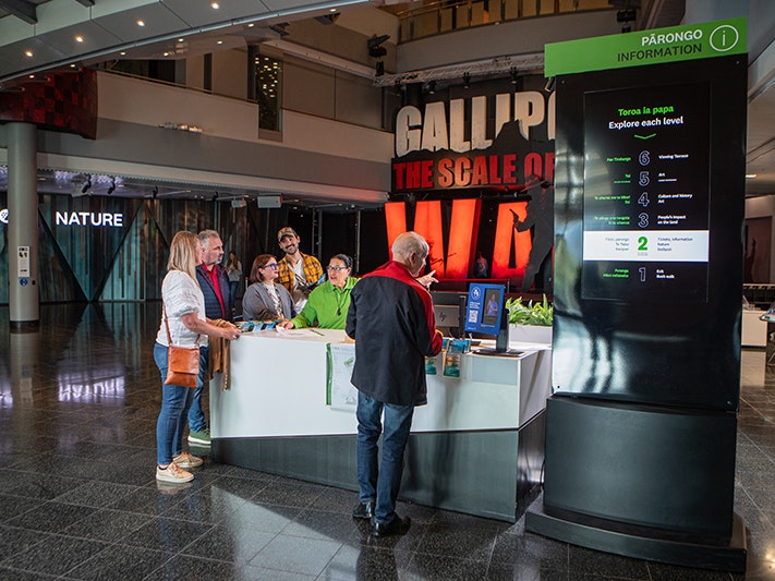 Te Papa information desk, 2025. Photo by Jeff McEwan. Te Papa (267091) Several people are standing by a desk in a museum where a staff member is telling them information.