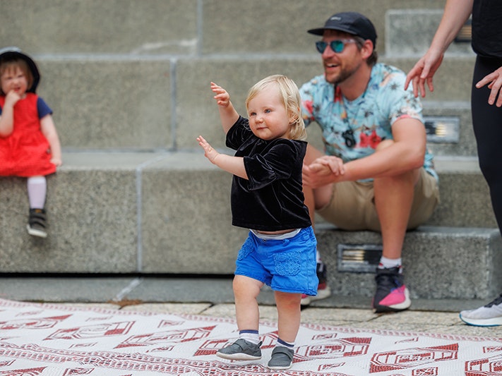 Raumati Whanau Day 2025, Photo by Jo Moore. Te Papa (257072) A small toddler is dancing on a mat while another toddler and two adults look on.