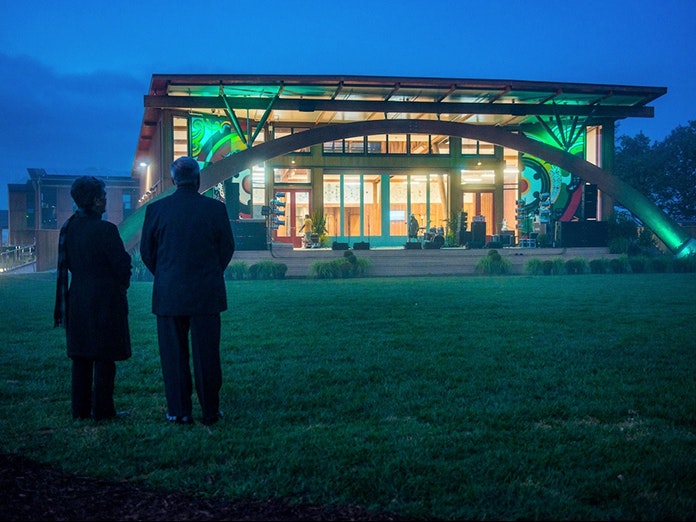 Still from Ever the Land. Courtesy of Sarah Grohnert Two people with their backs to the camera are looking at a lit-up building in a grass field at twighlight.