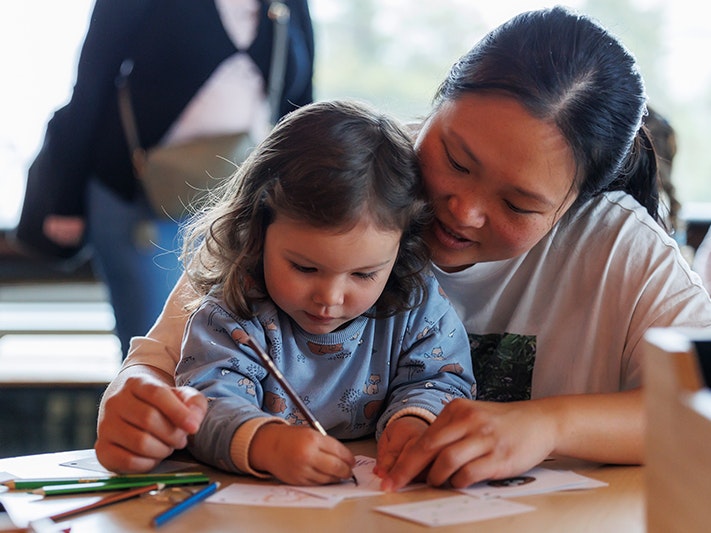 Whānau Day, 2023. Image by Jo Moore. Te Papa (228323) An adult and child working at a table with coloured pencils and paper.