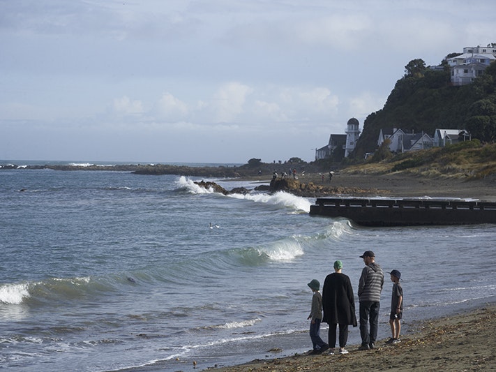 Two adults and two children are walking along a beach. There are some houses perched on a hill in the background.
