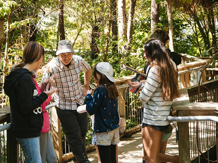 Curator Botany Leon Perrie at Whānau Day with DOC, 2020. Photo by Abbie Dorrington. Te Papa (162348) A smiling Leon in a bucket hat stands on a bridge in Bush City. He is talking to a group of 5 children holding magnifying glasses and clipboards.