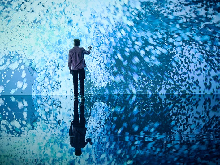 Marshmallow Laser Feast: Works of Nature, 2023, ACMI. Photo © Eugene Hyland Man standing in front of immersive installation of swirling blue light witha hand raised to touch it.