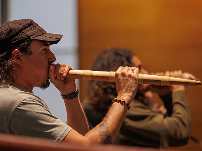 Curtis from Mauri Aura at After Dark: Mātahi o Rongo 2025. Photo by Jo Moore. Te Papa (261819) A man with long hair, a beard and cap is in the foreground playing a pūkāea - a long, narrow traditional Māori taonga puoro instrument, used to welcome people. A second man plays a taonga puoro instrument behind him.
