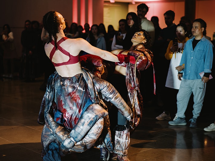 Body Island performing at Auckland Art Gallery, October 2025. Photo by David St George. Courtesy of Auckland Art Gallery Toi o Tāmaki Two performers wearing costumes like puffy exoskeletons hold hands as they lean back. Viewers stand around them, watching on.
