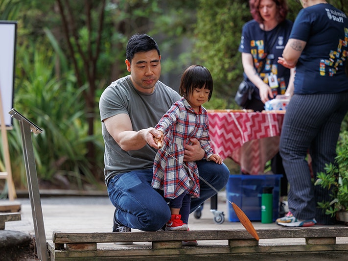 Raumati Whānau Day 2025. Photo by Jo Moore. Te Papa (257122) Man with little girl playing and whirling a wooden instrument outside.