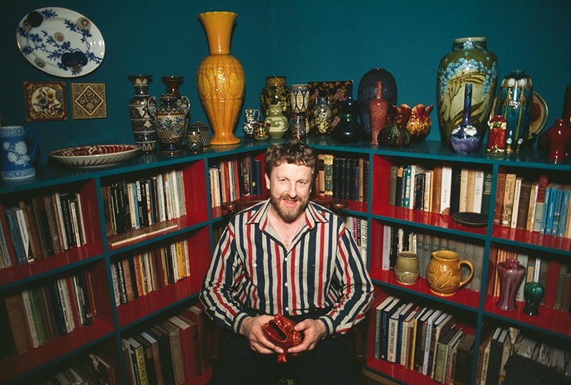 Portrait of Walter Cook, date unknown. Courtesy of Walter Cook A man in a stripey shirt and beard is smiling and holding a pot in front of a collection of books and pots.