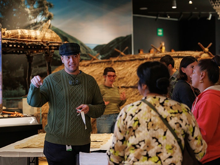 Mātauranga Māori Curator Isaac Te Awa. After Dark: Mātahi o Rongo 2025, photo by Jo Moore. Te Papa (261982) A male Curator is standing in an exhibition facing the camera, wearing a flat cap, green sweatshirt and a hei tiki pounamu taonga around his neck. He speaks to two women who have their back to the camera, holding a small piece of material in his hand.