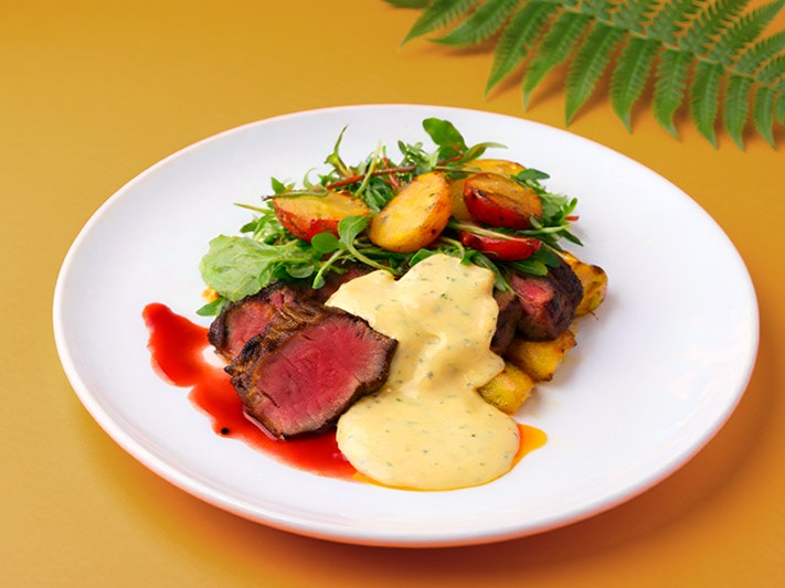 Te Papa Cafe Raumati Specials 2026, featuring seasonal dishes. Photo by Caitlin Dorrington. Te Papa (276290) A plate on an orange table with sirloin steak, green salad, potatoes and a creamy sauce. There is a green fern in the top right-hand corner of the table.