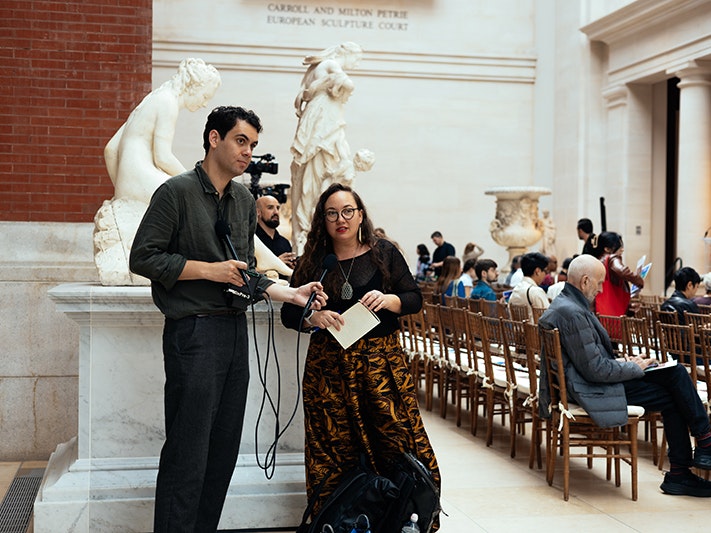 Jamie Tahana and Matariki Williams. Photo by Taylor Galmiche A tall man and a shorter woman are standing in front of a marble statue of a person sitting. Behind them are several people sitting in rows of chairs.