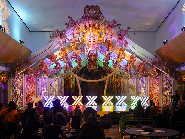 Matariki After Dark 2024. Photo by Neil Price. Te Papa (245519) A room with coloured lighting and colourful carving behind the stage. On the stage are nine lights in x shapes.