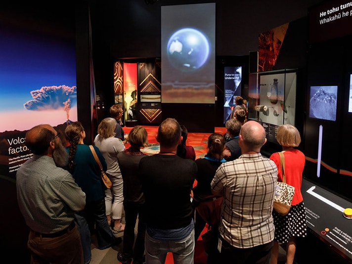 Meet the Scientists Teacher PD, 2021. Photo by Jess Dewsnap. Te Papa (168294) Several people standing in a museum space looking at a screen with an image of the world on it, with NZ showing.