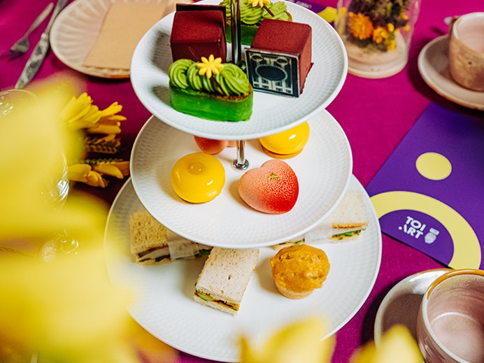 Eat Your Art Out high tea at Te Papa, 2026. Photo by Beeline Creative. Te Papa (279566) A set of three plates on a stand with different food on each plate, surrounded by a colourful tablecloth and cups and saucers.