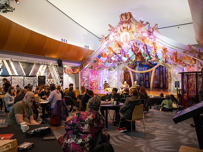 Matariki After Dark, 2025. Photo by Mel Waite, Te Papa (266412) People sitting at tables and on the floor in front of three seated speakers on the raised platform in front of the wharenui at Te Papa.