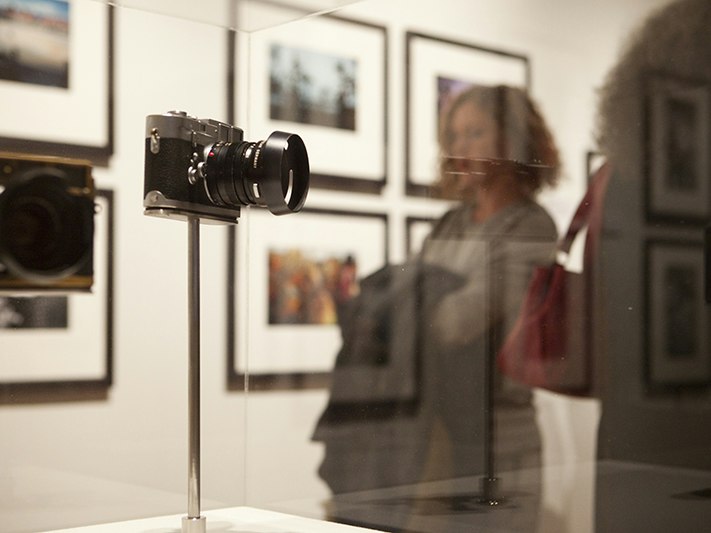 Brian Brake Lens on the World exhibition opening. Photo by Kate Whitley. Te Papa (55689) A person is out of focus behind two cameras in a glass stand in a gallery.