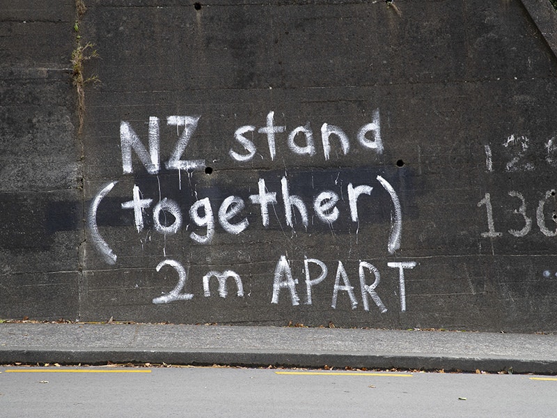 Graffiti on a concrete wall beside a road. It says "Stand Together (2m apart)"