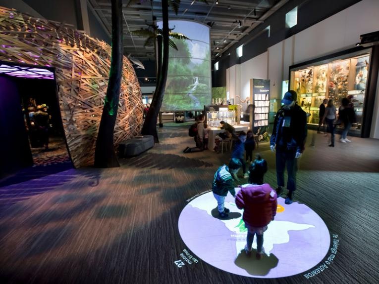 Children are standing on a projected map of the world whila an adult looks on.