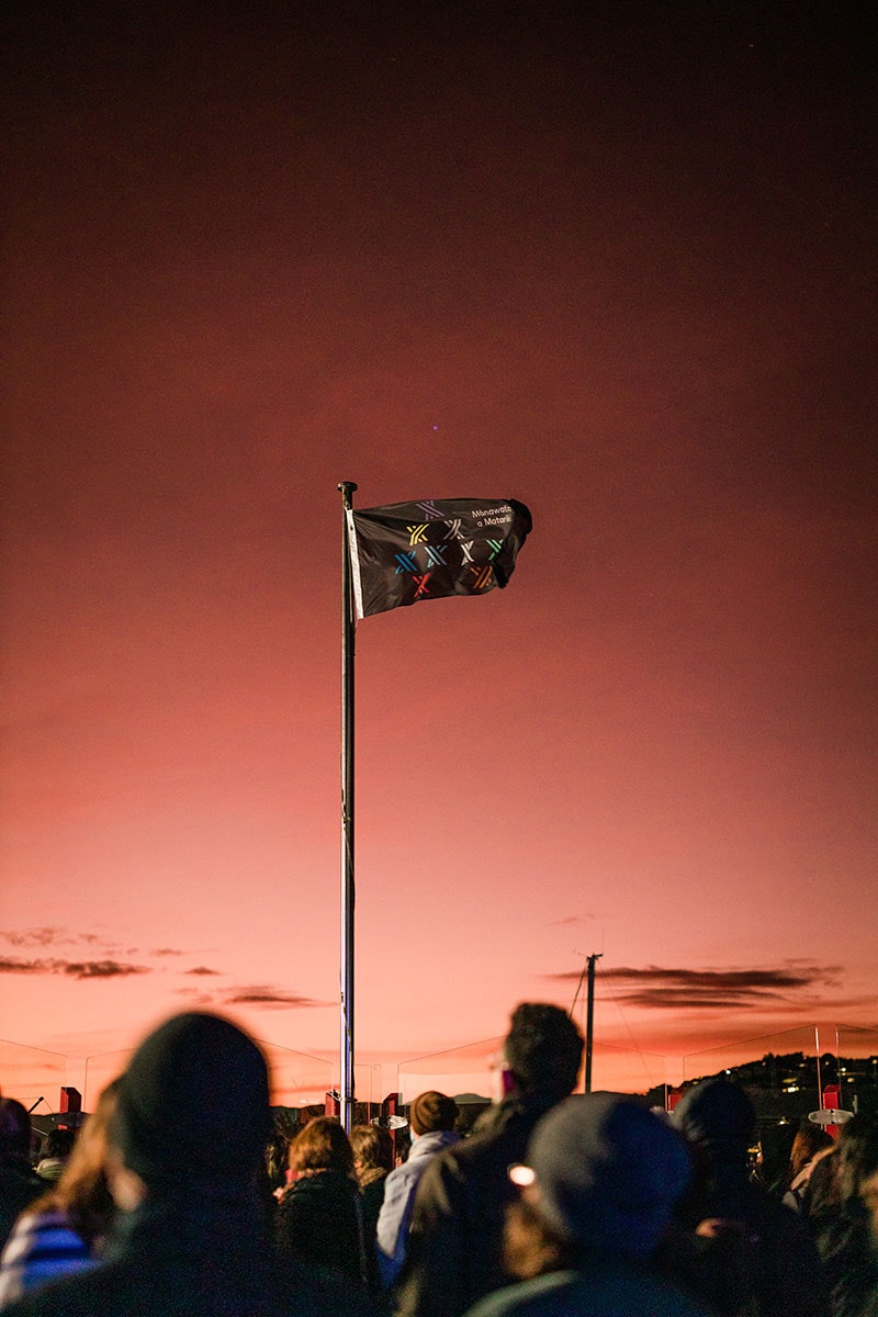 A black flag with the logo of the Matariki public holiday, consisting of nine stylised stars, flies in the early morning Wellington breeze, with an orange sunrise behind it