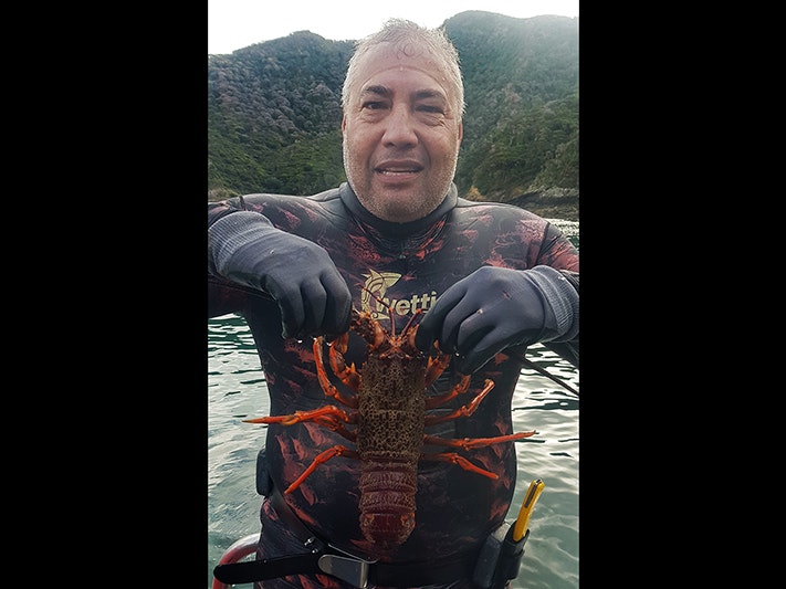 Tamati in dive gear holding a crayfish. Photo courtesy of Tamati Stevens A man in a wetsuit is holding a crayfish up in front of himself. He is looking at the camera.