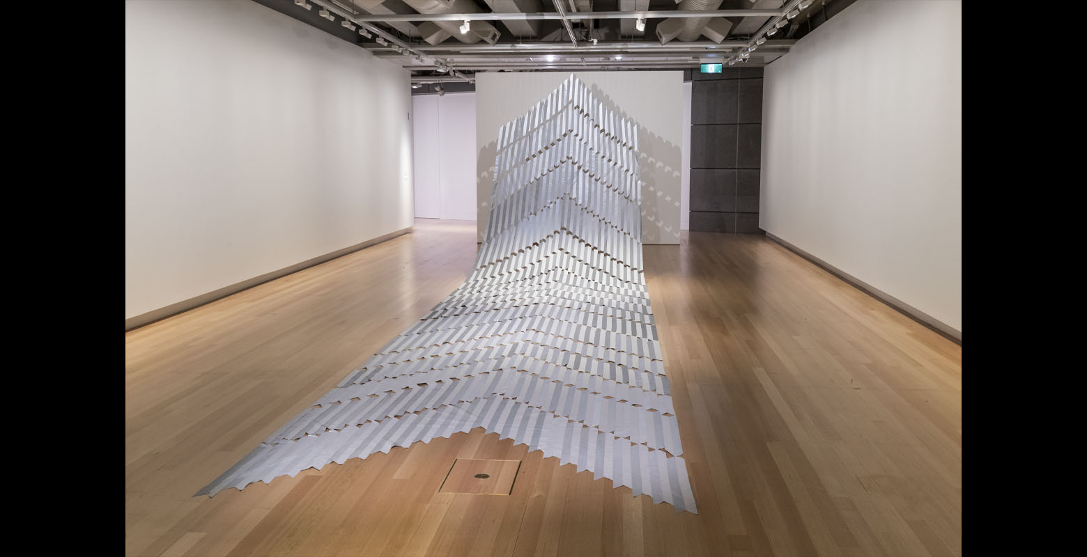 Pieces of hi-vis tape are woven together to create a large arrow-shaped artwork point from the floor up to the ceiling.
