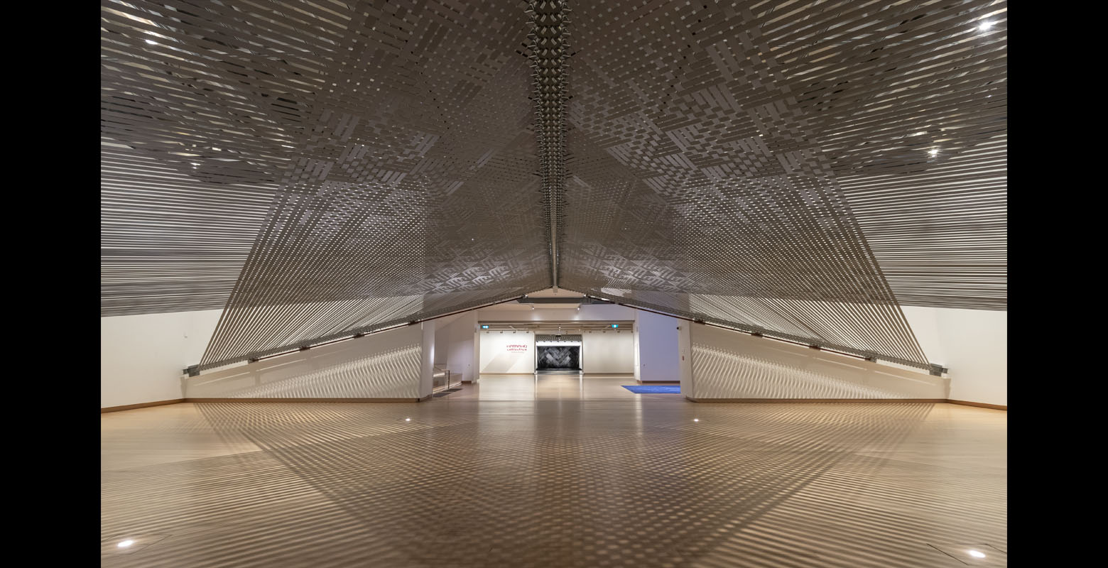 A large-scale, woven installation that is suspended from above and spans a double-height gallery space with a wooden floor and white walls. Woven, grey and reflective tie-downs create shadows and reflections on the walls and floor.