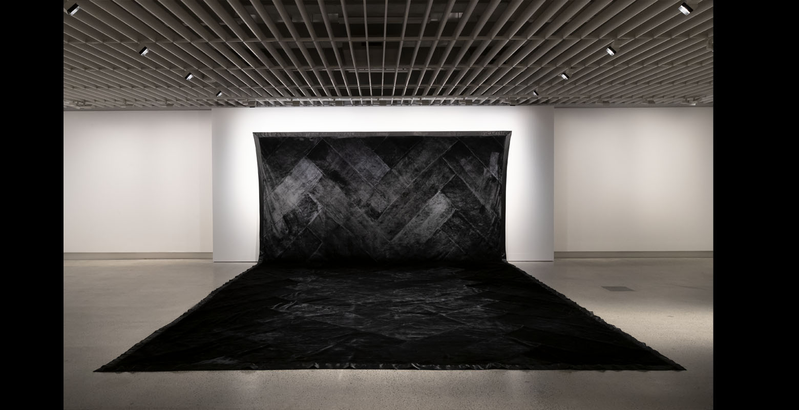 Multiple black blankets are sewn together to form a giant blanket, which hang from a wall and drapes over the floor. The blankets are stitched together diagonally, forming mountain-like patterns.