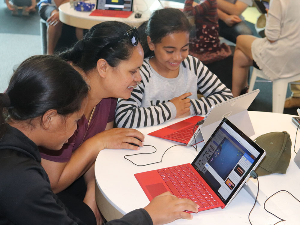 A woman and two young girls sit at a desk, coding on computers