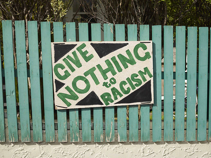 Green fence with a hand-painted sign on it saying, 'Give nothing to racism' in green and black.