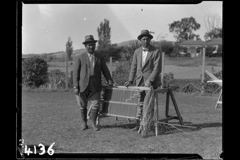 Black and white photo of two people standing in a field next to a piece of weaving