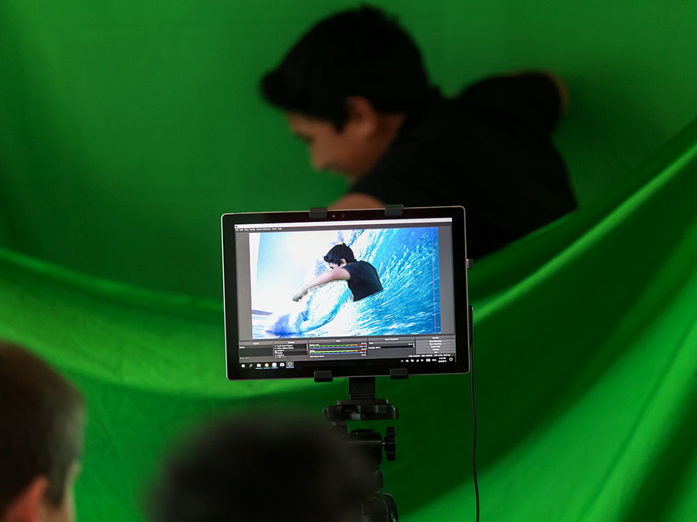 Two children play with green screen. You can see one child through the screen of a camera, acting in front of a green screen