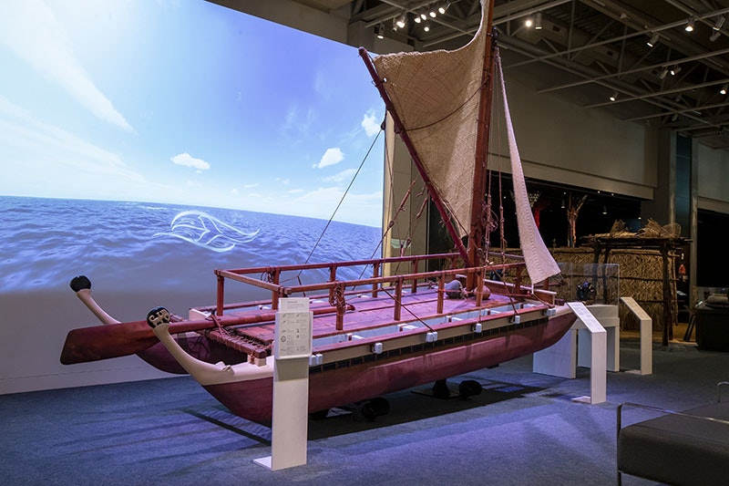General view of an exhibition space with a large projection on the wall, a waka (Māori canoe) in the foreground, and further exhibits in the background