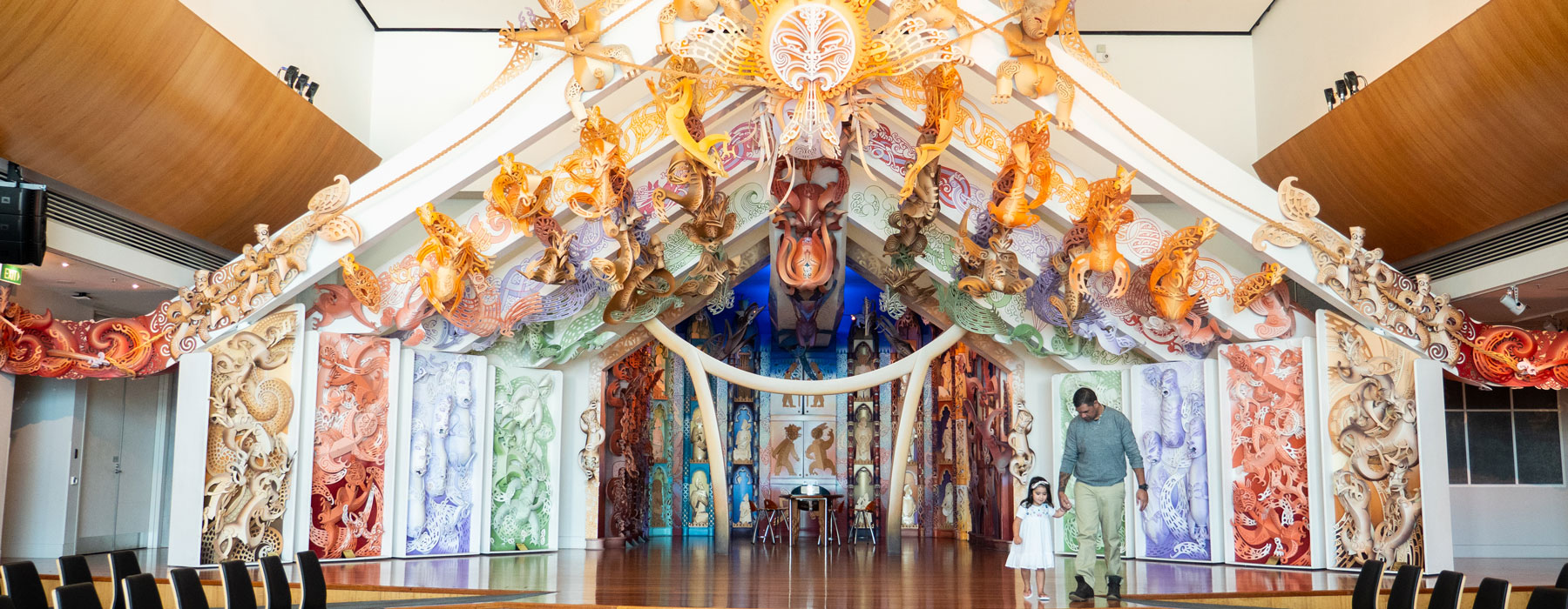 A father and daughter hold hand on an ornately carved wharenui (large meeting house)