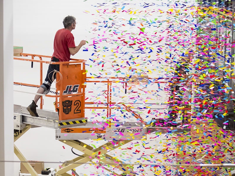 A man stands on a scissor lift hanging strands of coloured plastic from the ceiling while a woman below holds the strand so it doesn't get tangled