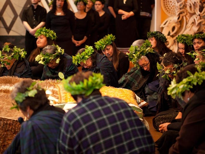Several women wearing black and wreaths on their heads made of kawakawa leaves are sitting around a cloak on some boxes in front of them.