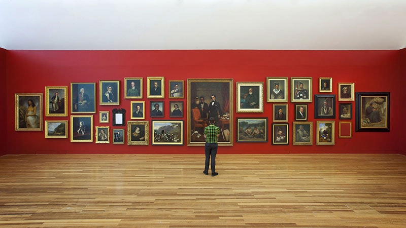 A man stands looking at a red wall featuring paintings hung in the salon style