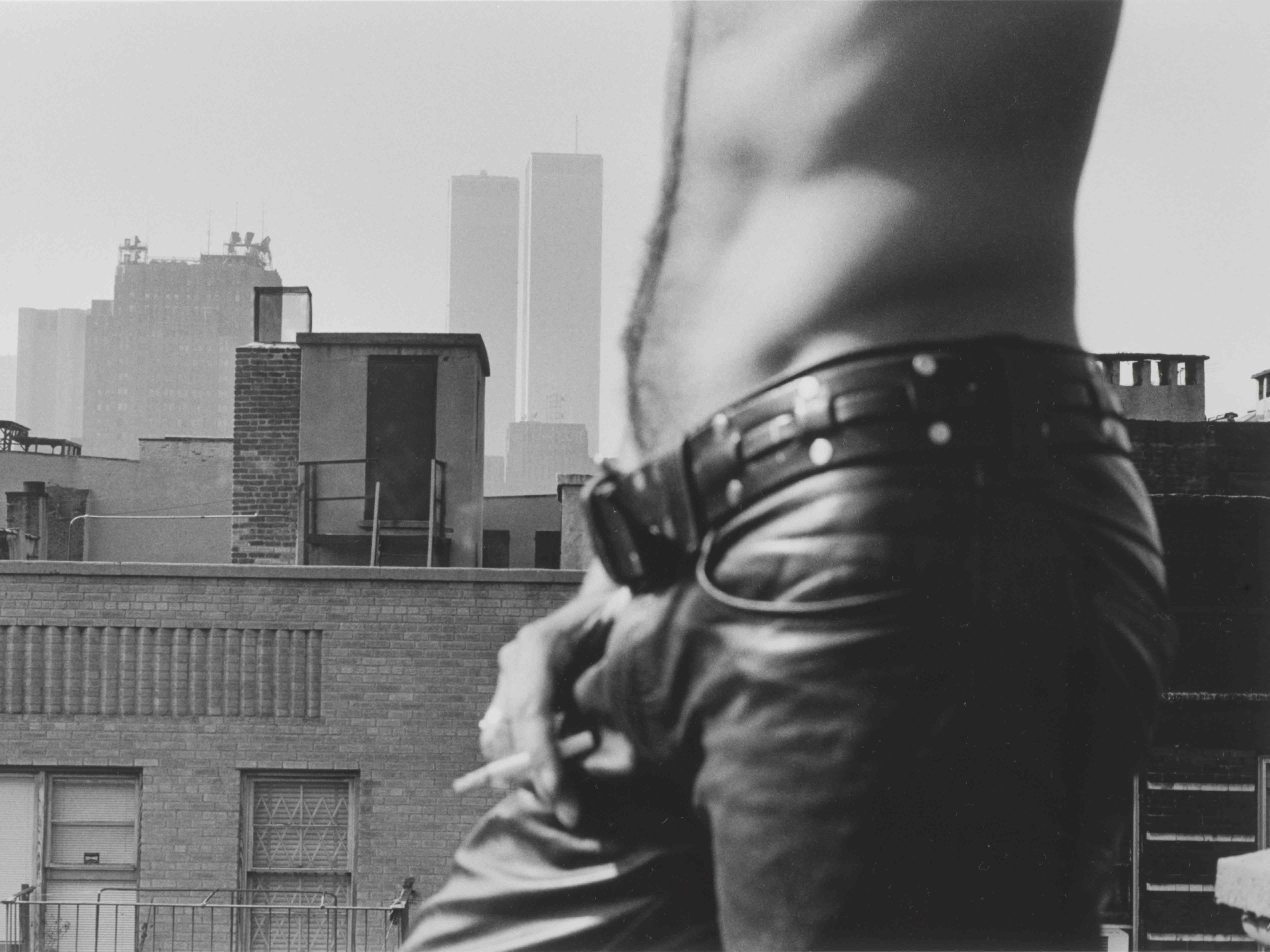 A black and white photograph of a man's torso. He is wearing jeans and smoking, standing on a roof with a city in the background.