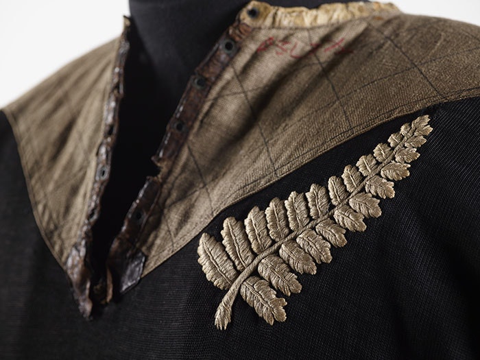 Close-up of 1905 All Blacks jersey, featuring a brown crest on the top and a large silver fern on the left-hand side. The jersey is black