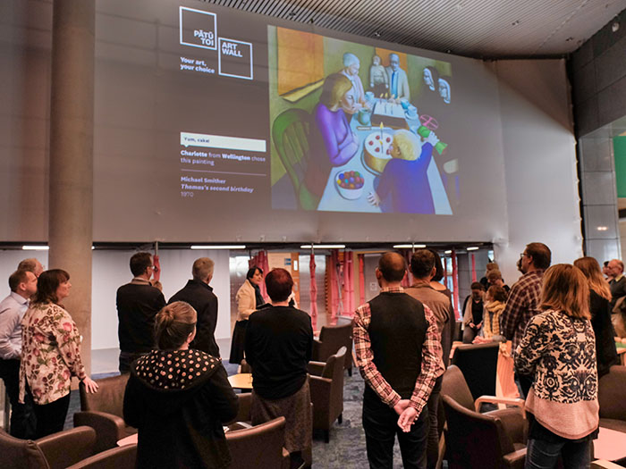 A group of people listen to a person talk while standing in front of an art work being projected onto a wall