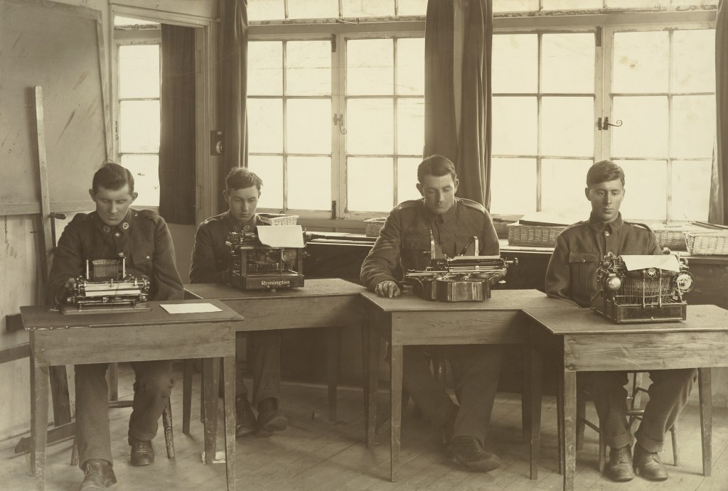 Three soldiers missing an arm each sit in a classroom in front of typewriters
