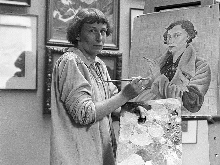 Rita Angus looks towards the photographer in a portrait of her painting herself