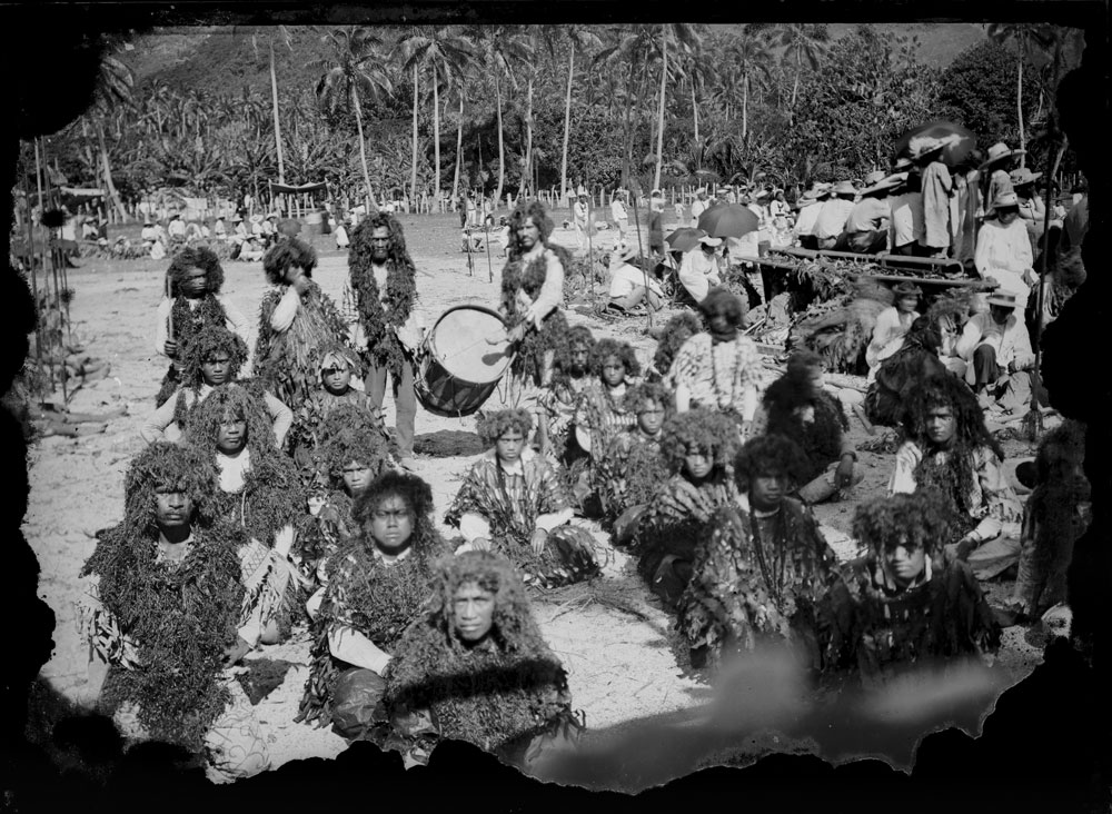 Group of performers dressed in elaborate costumes with a man standing behind them beating a drum. In the background are rows of coconut palms
