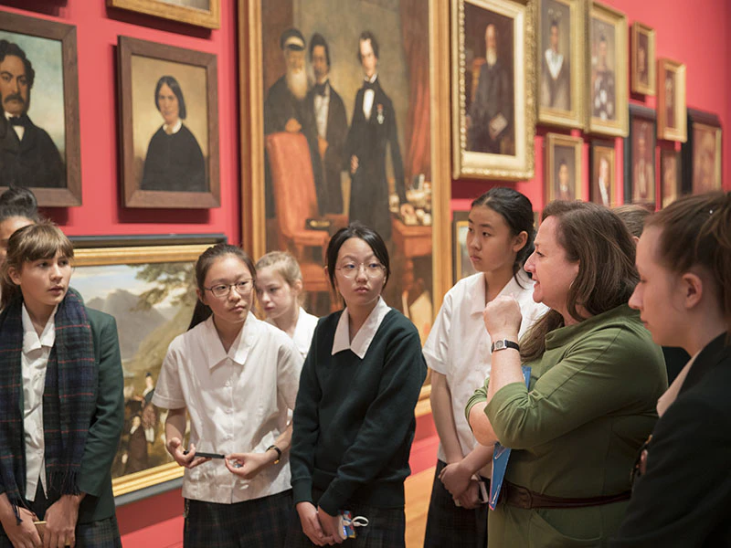Students and a teacher stand in front of a wall of paintings