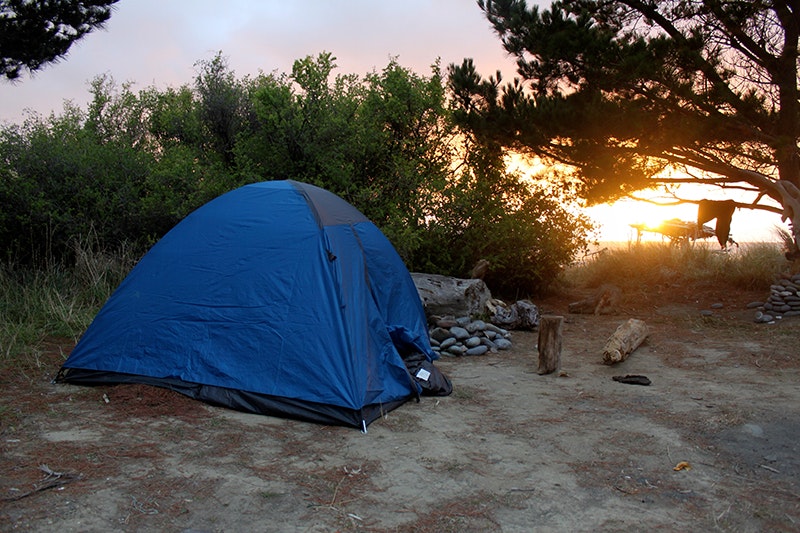 A blue tent set up for camping in a tree-lined area. There is a sunset in the background.